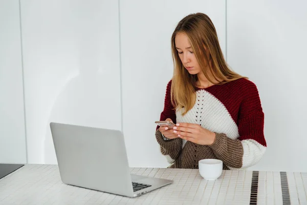 Chica bonita mirando la pantalla del teléfono mientras usa el ordenador portátil, en casa — Foto de Stock