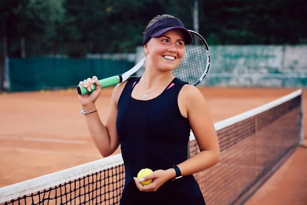 Tennis player. Happy girl, standing with racket and tennis ball on the court, near the net, smiling widely, looking away. Sport concept.
