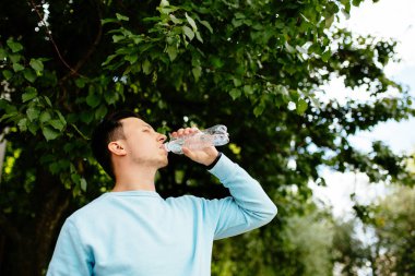 Genç adam yaz aylarında yeşil ağacın arka planında şişeden su içiyor