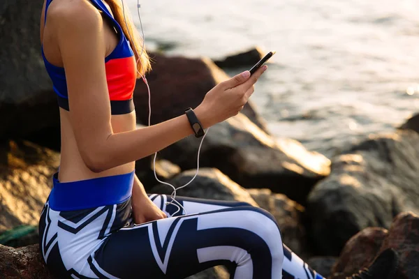 Mujer deportiva usando un smartphone, sentada cerca del océano — Foto de Stock