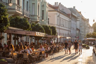 Szeged, Macaristan - 2 Temmuz 2018: tablolarda kafe ve restoranlarda oturan diğer kişilerle Szeged, Güney Macaristan, bir yaya caddesi üzerinde yürüyen genç insanlar