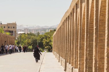 ISFAHAN, IRAN - AUGUST 7, 2015: iranian woman wearing the traditional muslim black veil waling on Si o Seh Pol bridge. Also known as Allahverdi Khan Bridge,  it is a major landmark of the city clipart