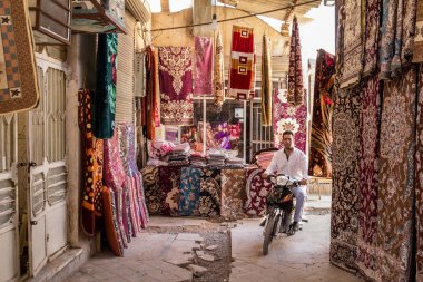 YAZD, IRAN - AUGUST 18, 2015: Man driving a motorcycle surrounded by stores selling carpets & rugs in Yazd Khan bazaar. Yazd is one of the main cities of central Iran clipart
