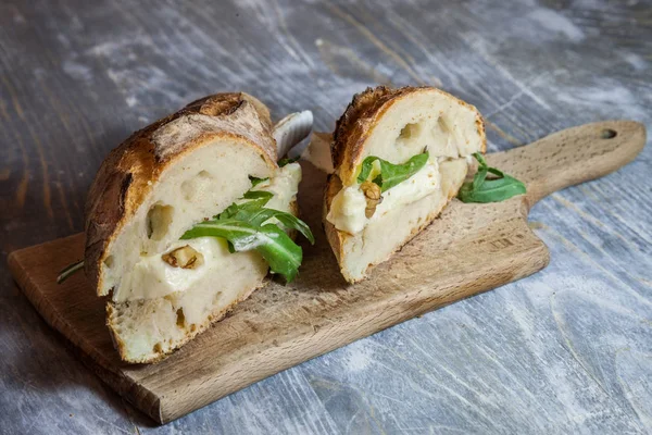 Brie sanwdich in a French baguette, made of Brie de Meaux Cheese with some slices of rucola salad and walnuts on display on a rustic wooden table. This sandwich, in France, is called sandwich au brie