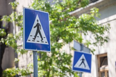 İki yol işareti, şehir manzarasında yayaların geçtiği bir yaya geçidinin varlığına dair uyarıda bulunuyor. Avrupa standartlarına uygun, tipik mavi şablonuyla.