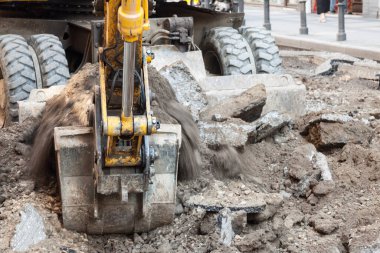 Focus on gravel and dirt going out of the showel of a mechanical digger excavator working in a construction site of a street renovation urban program clipart