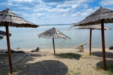 Üç yıpranmış kamış şemsiyesi, Sırbistan 'ın Tuna Nehri boyunca uzanan dar bir çakıl taşı plajına, sakin mavi sulara ve dağınık yaz bulutlarının altında ağaçlarla kaplı uzak bir kıyıya dökülüyor..
