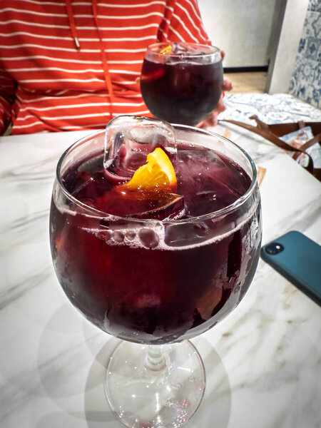 Close-up of two glasses of red sangria with ice cubes and orange wedges on a marble table in a Valencia bar. Condensation glistens on glass while a background striped sweater suggests relaxed social atmosphere.