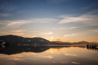 Klagenfurt, Avusturya 'da Worthersee üzerinde gün batımı. Altın gökyüzü göl yüzeyine yansıdıkça dağlar karanlık siluetler oluşturur. Alacakaranlıkta sakin panoramik bir su manzarası.