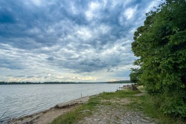 Sırbistan 'ın Grocka kentindeki Tuna Nehri' nin geniş bir nehir yüzeyi üzerinde kıyı şeridi taşları, yeşillikler ve dramatik bir bulut güvertesi yer alıyor. Manzara Belgrad yakınlarındaki sakin bir nehir manzarasını taşıyor.