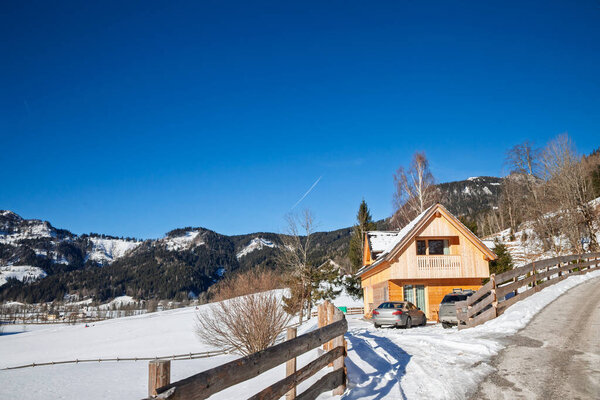 Деревянное альпийское шале в Словении в солнечный зимний день, с подъездной дорожкой, припаркованными автомобилями и огороженным участком с видом на снежные холмы, иллюстрирующее горную недвижимость в Альпах и современную жизнь в хижине