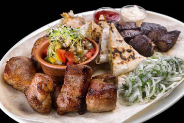 Karışık ızgara etler, kebaplar, domuz, sığır eti, tavuk. Pide, soğan ve ızgara sebzeler ile. Beyaz bir plaka üzerinde. Siyah bir arka plan üzerinde.