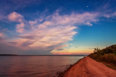Gölün üzerinde güzel bir günbatımı, bulutlar ve kıyı boyunca uzanan bir yol.