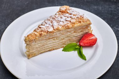 Bir parça kek Napolyon, beyaz bir tabakta, çilek ve nane ile, koyu bir arka planda