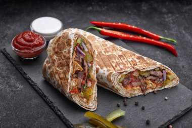 Shawarma soslu, soğanlı, turşulu, otlu ve acı kırmızı biberli, siyah beton arka planda.