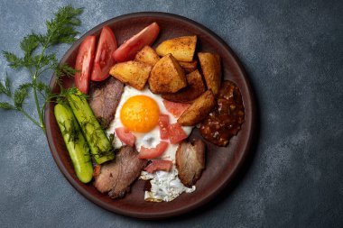 Kil tabakta kahvaltı, salatalık, kızarmış patates, sahanda yumurta, domuz pastırması, domates ve sos, otlarla birlikte mavi beton üzerine.