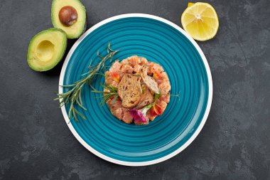 Avokadolu somon tartare, mavi çizgili tabakta, kireç ve kızarmış ekmekle, koyu bir arka planda.