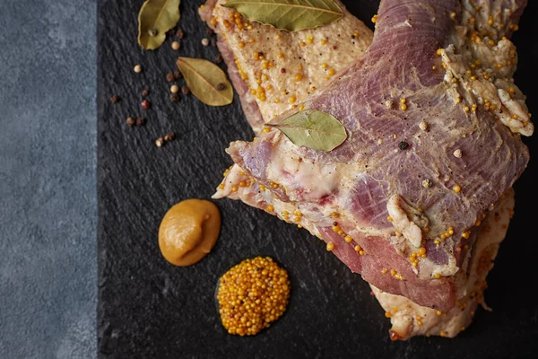 Two slices of raw pickled pork underscore, with mustard, pepper and bay leaf, against a dark background