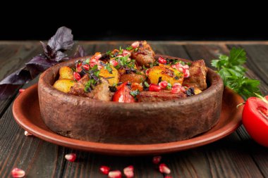 Et ve nar tohumlu kızarmış patates, kil bir tabakta, ahşap bir arka planda otlar ve domatesle.