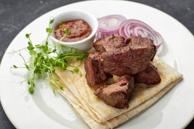 Lavaş, sos, mikroyeşil ve soğanlı kızarmış şiş kebap, beyaz bir tabakta, gri bir arka planda.