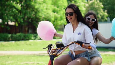 İki genç seksi esmer bağsız kıl ile kısa denim şort ve beyaz t-shirt parkta gülüyor ve tatlı pamuk ile gülümseyen bir elektrikli motosiklet devam