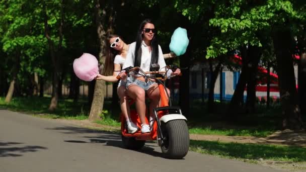 Deux jeunes et sexy brune incendiaire aux cheveux lâches en short denim et chemises blanches vont sur une moto électrique dans le parc, s'embrassant — Video