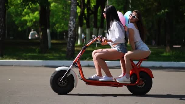 Две юные и сексуальные брюнетки с распущенными волосами в коротких джинсовых шортах катаются на электрическом мотоцикле в парке в солнечный день, наслаждаясь объятиями друг друга. Лучшие друзья проводят время вместе — стоковое видео