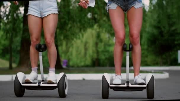 Dos chicas jóvenes sexy cabalgando en Segway en pantalones cortos tomados de la mano y riendo en un día soleado en el parque — Vídeos de Stock