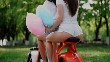 İki genç ve seksi esmer arkadaşı gevşek saç ile kısacası parkta bir elektrikli motosiklet Binme kot şort. Yakın çekim manzara arka yüzü. Güneşli bir günde bir scooter güzel kalça