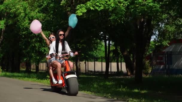 Duas morenas sensuais jovens com cabelo solto em shorts jeans curtos e camisetas brancas vão em uma motocicleta elétrica no parque rindo e sorrindo com algodão doce — Vídeo de Stock