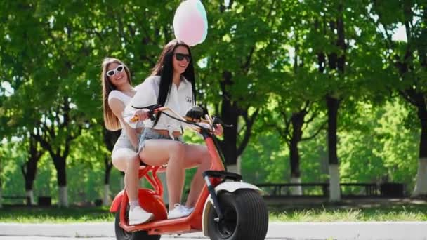 Dois jovens e sexy morena amigos com cabelo solto em shorts jeans curtos montando uma motocicleta elétrica no parque em um dia ensolarado gostando de abraçar uns aos outros. Melhores amigos passam tempo juntos — Vídeo de Stock