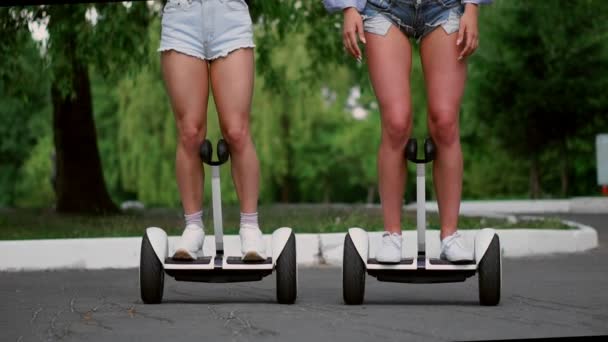 Dos chicas jóvenes sexy cabalgando en Segway en pantalones cortos tomados de la mano y riendo en un día soleado en el parque — Vídeos de Stock