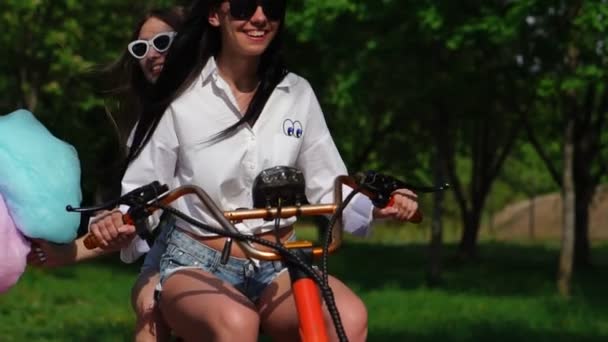 Twee jonge en sexy brandstichting brunette met losse haren in het kort denim shorts en witte shirts gaan op een elektrische motorfiets in het Park, genietend van knuffelen elkaar — Stockvideo