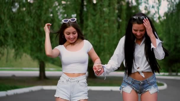 Zwei sexy junge Mädchen, die in kurzen Shorts auf dem Segway fahren, Händchen halten und an einem sonnigen Tag im Park lachen — Stockvideo