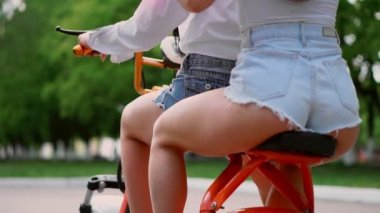 İki genç ve seksi esmer arkadaşı gevşek saç ile kısacası parkta bir elektrikli motosiklet Binme kot şort. Yakın çekim manzara arka yüzü. Güneşli bir günde bir scooter güzel kalça