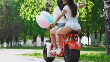 İki genç ve seksi esmer arkadaşı gevşek saç ile kısacası parkta bir elektrikli motosiklet Binme kot şort. Yakın çekim manzara arka yüzü. Güneşli bir günde bir scooter güzel kalça