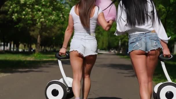 Two women in denim shorts and white t-shirts close - up go from the rear view camera and carry in the hands of a self-balancing scooter — Stock Video