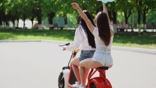 Dois jovens e sexy morena amigos com cabelo solto em shorts jeans curtos montando uma motocicleta elétrica no parque em um dia ensolarado gostando de abraçar uns aos outros. Melhores amigos passam tempo juntos — Vídeo de Stock