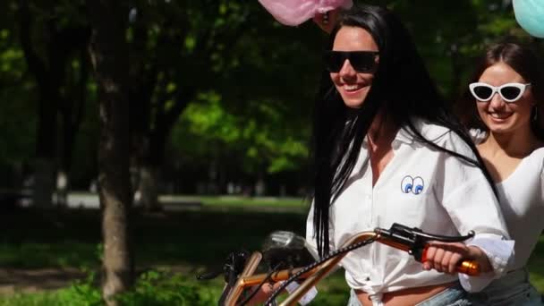 Две красивые девушки в джинсовых шортах летом едут в парк на электрическом скутере — стоковое видео