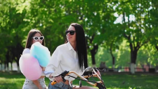 Две красивые девушки в джинсовых шортах летом едут в парк на электрическом скутере — стоковое видео