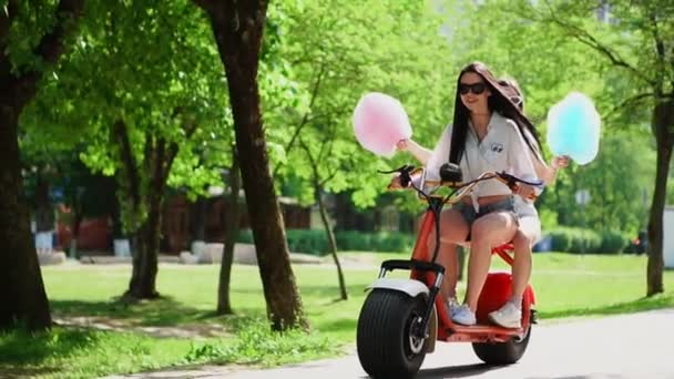 Två vackra flickor i denim shorts rida i parken på en elektrisk skoter på sommaren — Stockvideo