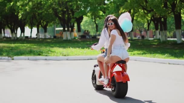 Dvě krásné dívky v džínové šortky jezdit v parku na elektrický skútr v létě — Stock video