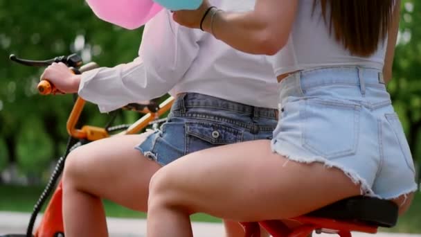 Dos hermosas chicas en pantalones cortos de mezclilla viajan en el parque en un scooter eléctrico en el verano en cámara lenta vista trasera — Vídeos de Stock