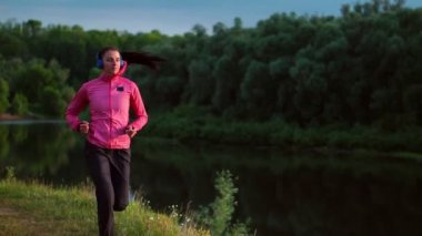 Kulaklık için maraton hazırlanıyor Nehri yakınında bir kız pembe bir ceket ve siyah pantolon çalışır