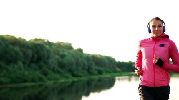 A girl in a pink jacket and black pants runs near the river in headphones preparing for the marathon — Stock Video