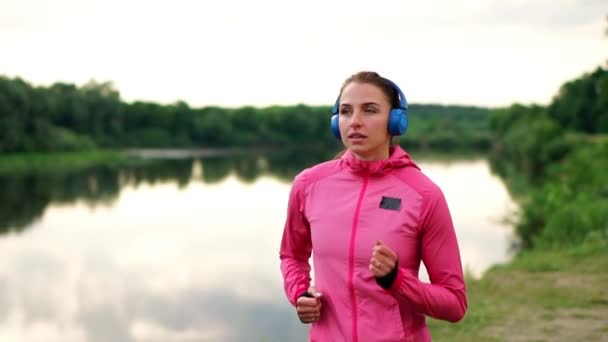 En flicka i en rosa jacka och svarta byxor kör nära floden i hörlurar förbereder för maraton — Stockvideo
