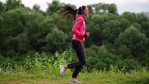 Dziewczyna w różowym kurtkę i czarne spodnie biegnie w pobliżu rzeki w słuchawkach przygotowania do maratonu — Wideo stockowe