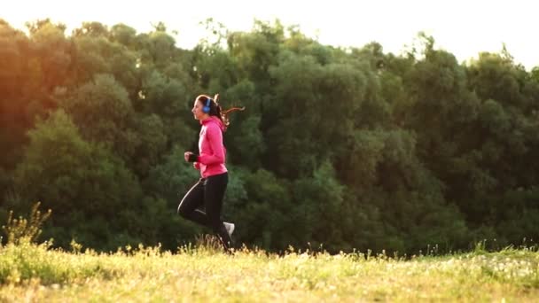 La chica corre al atardecer en el parque a lo largo del estanque y escucha música en los auriculares — Vídeos de Stock