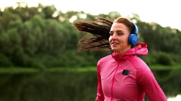 Een meisje in een roze jas en zwarte broek loopt in de buurt van de rivier in hoofdtelefoons voorbereiding voor de marathon — Stockvideo