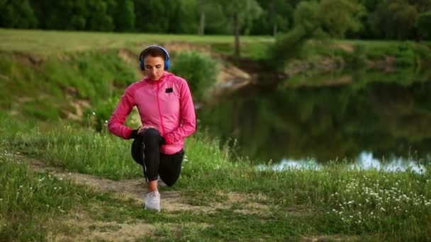 Una chica con una chaqueta rosa se está preparando para un calentamiento y escuchar música en los auriculares a través del teléfono — Vídeos de Stock